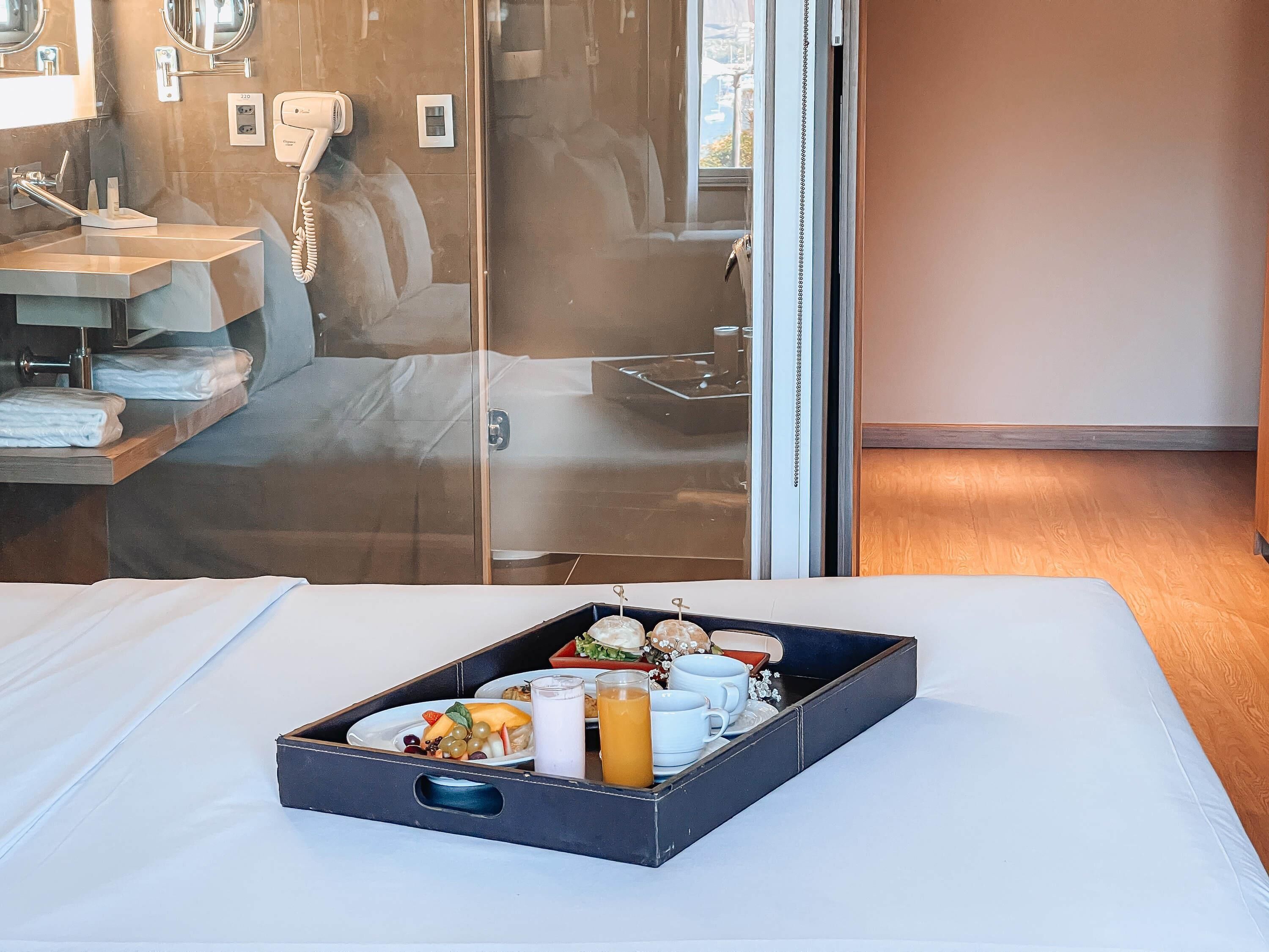 Habitación de hotel con cama, mesita de noche con lámpara, separador de vidrio, baño con espejo y bandeja con alimentos para el desayuno.