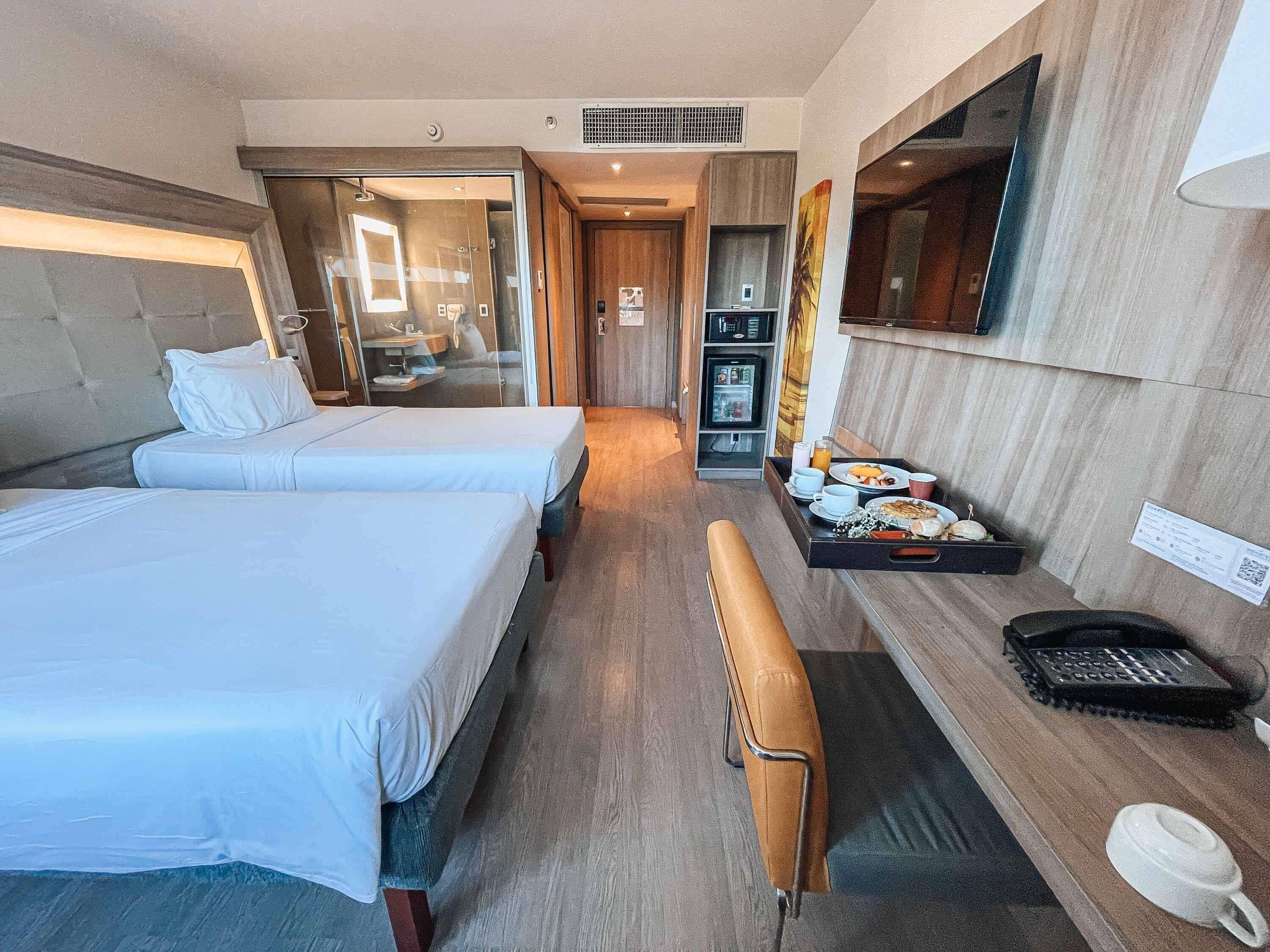 A hotel room with two beds, a bench, a desk with a telephone, a refrigerator, and a television.