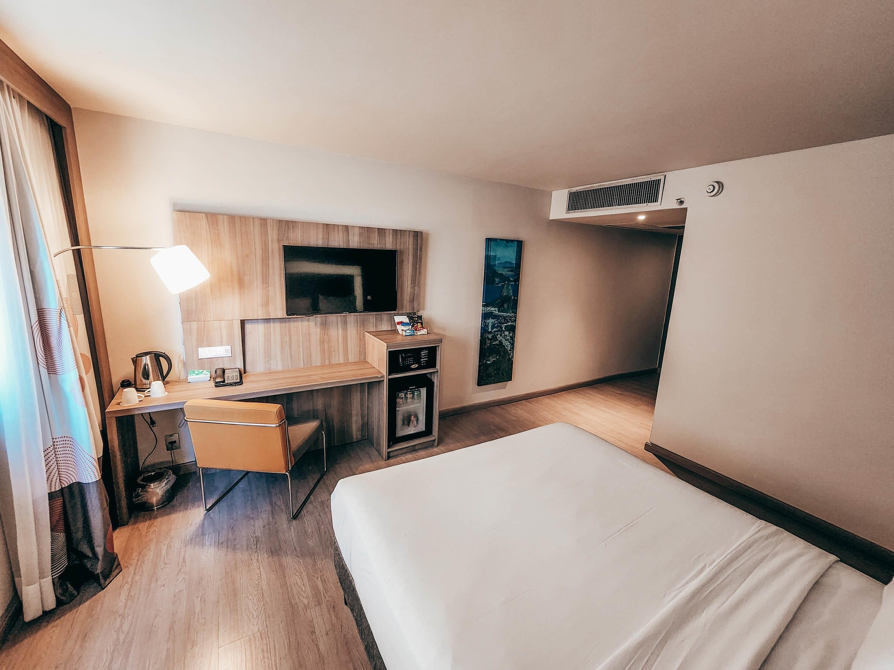Ett hotellrum med en säng, ett skrivbord, en stol, en TV och en minibar.