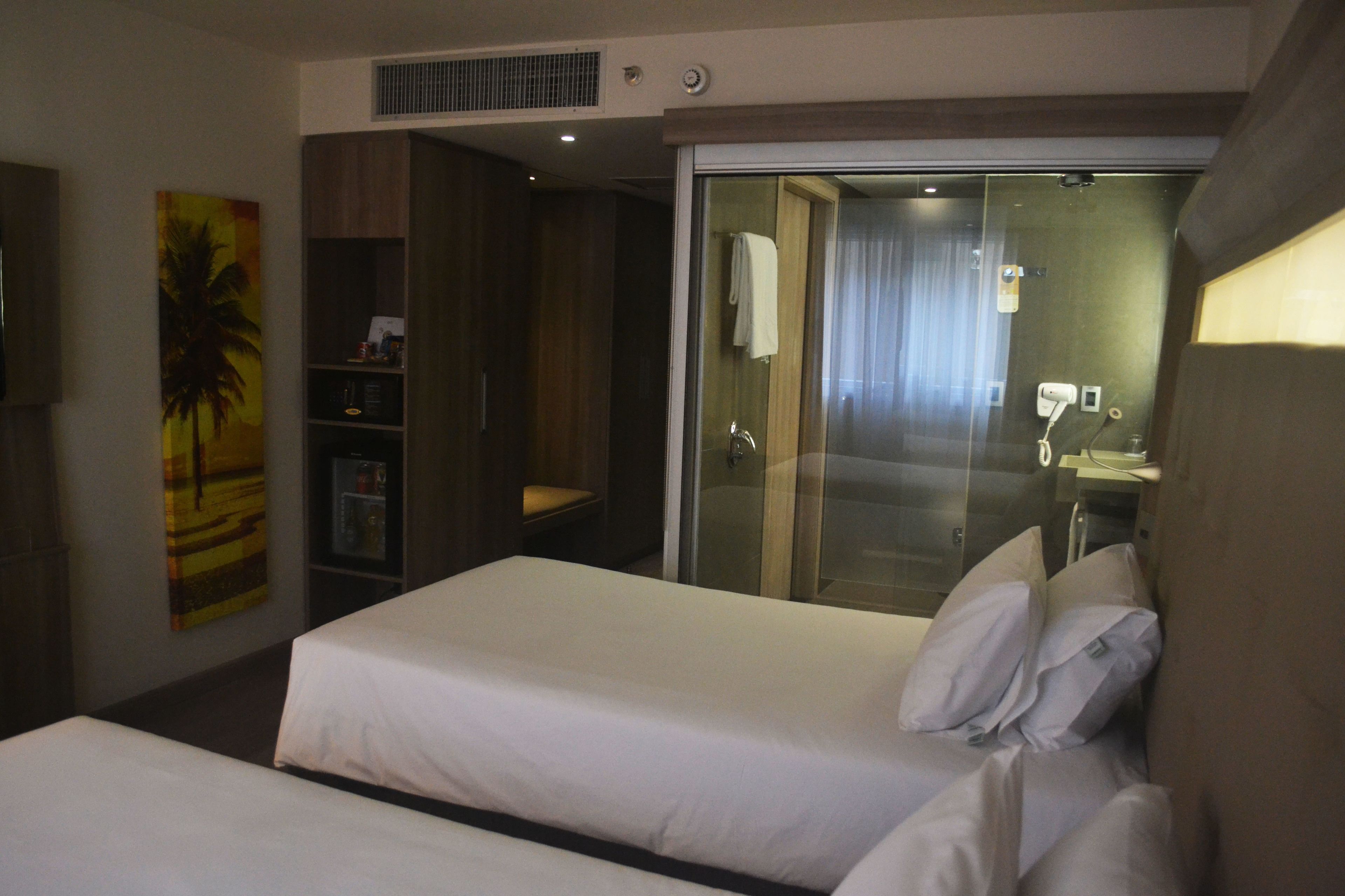A hotel room with two beds, a sliding glass door leading to a bathroom, and a wall-mounted TV.