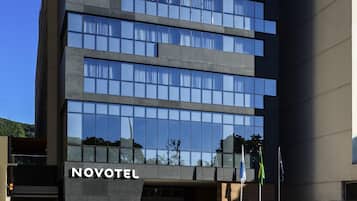 A modern hotel building with a clear glass facade reflecting the sky and trees. The name "NOVOTEL" is prominently displayed on the entrance.
