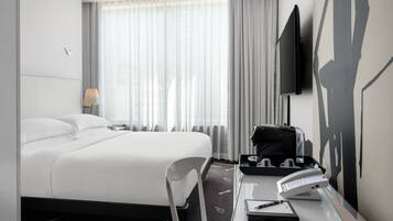 A modern hotel room with a large bed, a desk with a telephone and a notepad, a chair, a lamp, and a TV mounted on the wall.