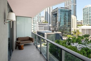 A balcony with a sofa, a small table, and a lamp. It overlooks a cityscape with modern buildings and a street below.