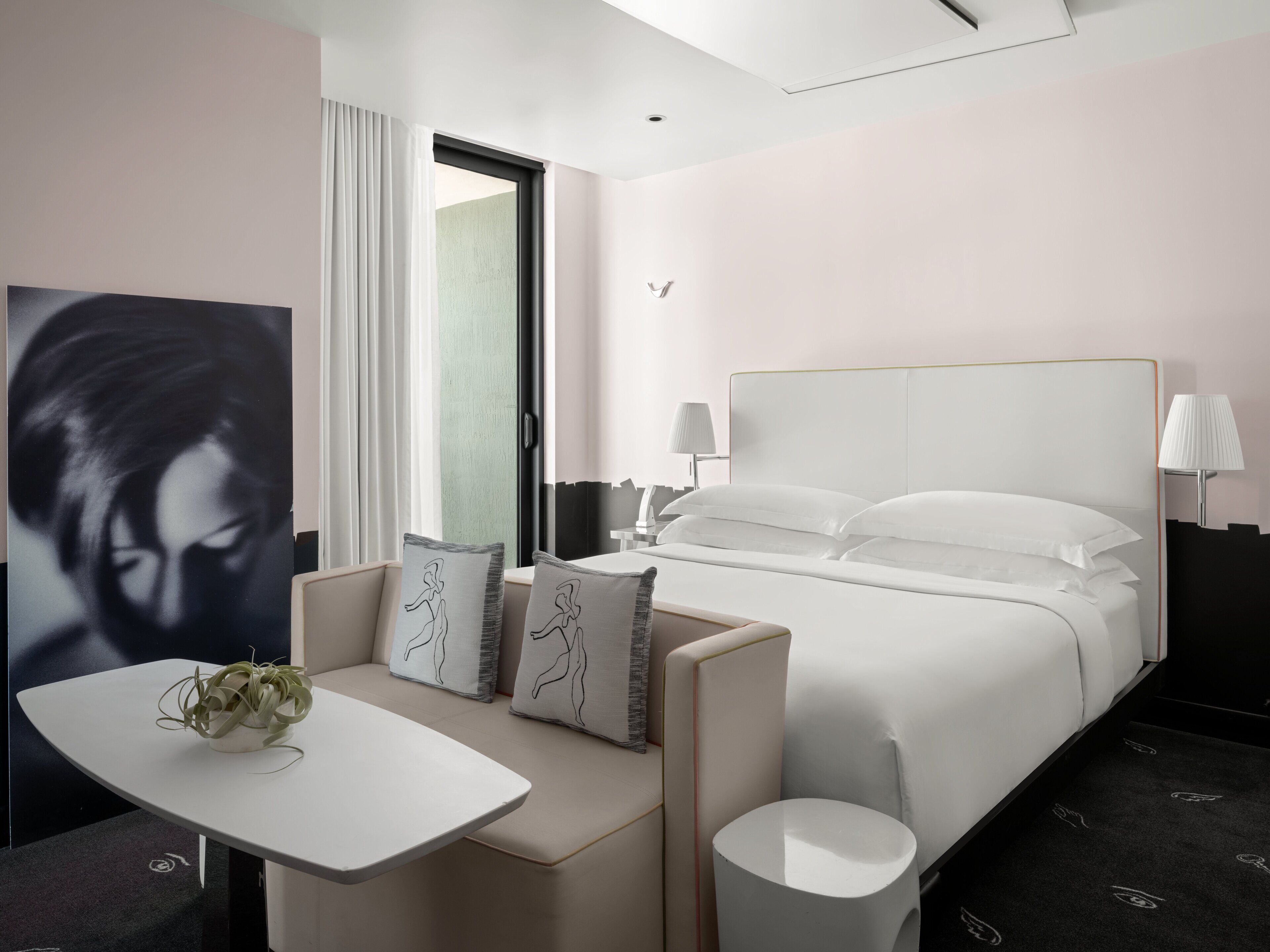 A modern hotel room with a bed, bedside tables, a small seating area, and a large artwork on the wall.