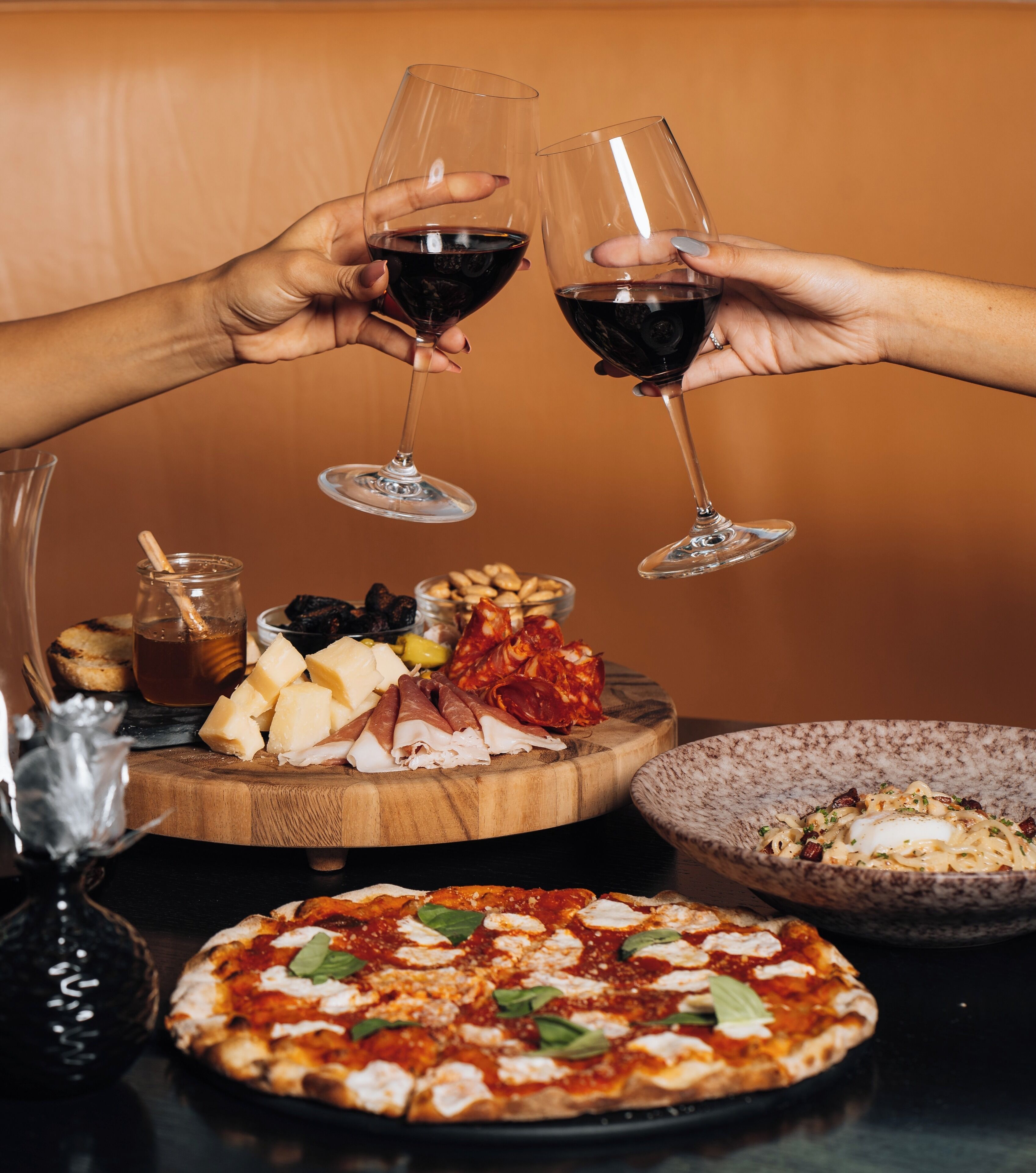 Pöydällä on pizzaa, juustoa, erilaisia leikkeleitä ja pullo viiniä.