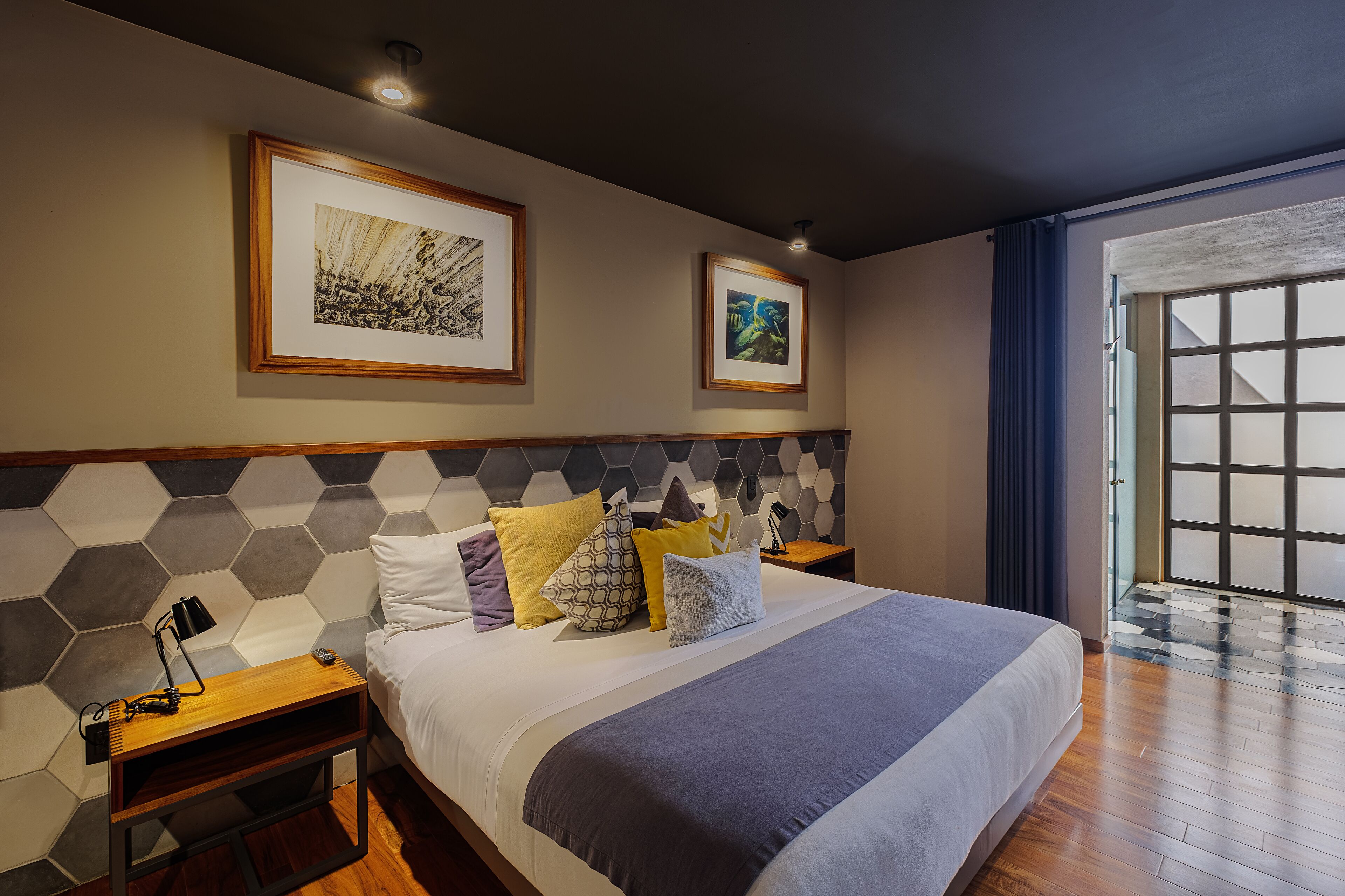 Quarto de hotel moderno com uma cama grande, cabeceira de madeira, duas obras de arte emolduradas, mesa de cabeceira com abajur e vista para o exterior por uma porta deslizante.