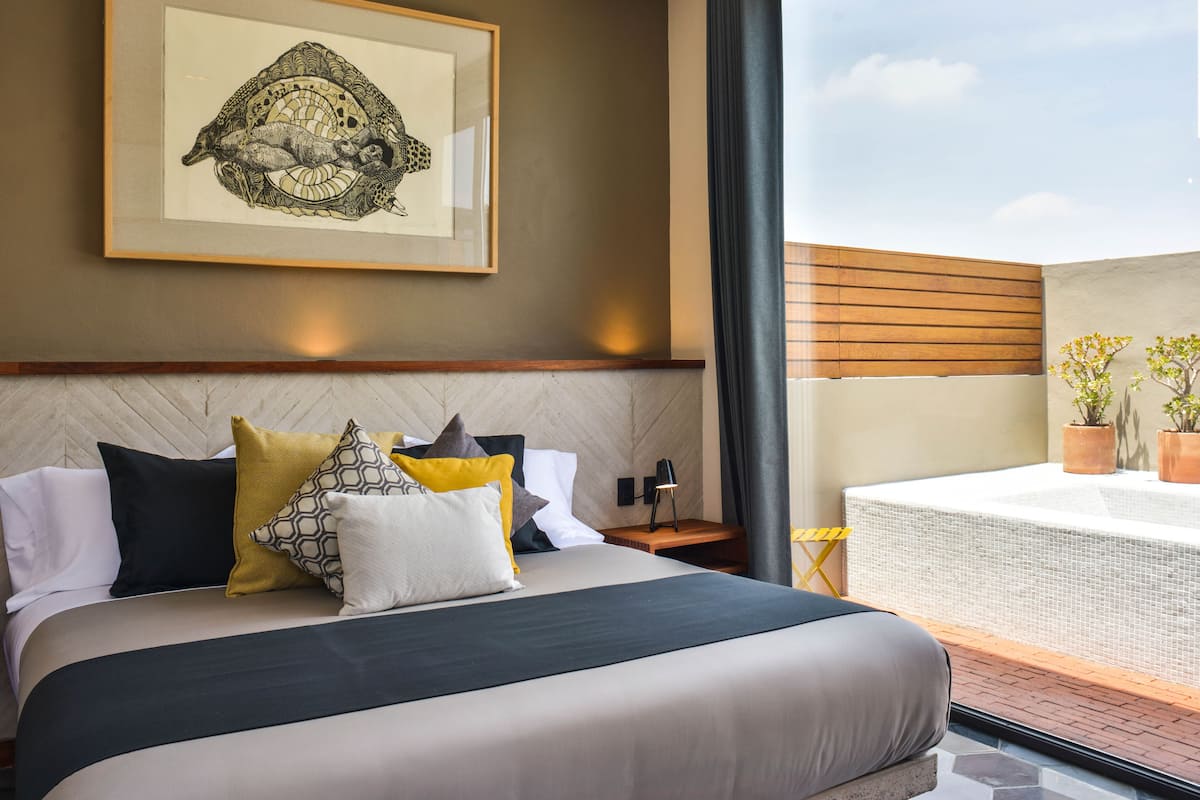 A modern hotel room with a large bed, a framed artwork above the bed, a balcony with a table and potted plants, and a view of the sky.