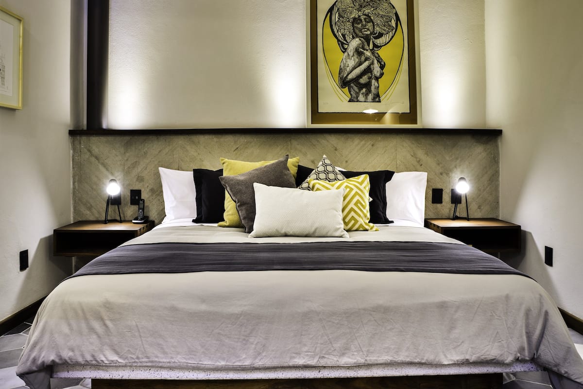 A modern hotel room with a large bed, multiple pillows, a headboard with artwork, bedside lamps, and a geometric-patterned floor.