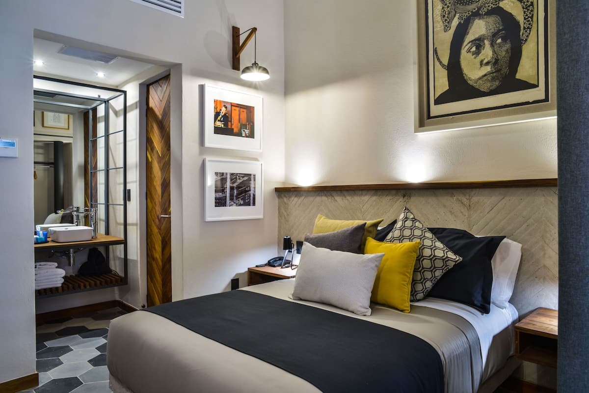 A modern hotel room with a large bed, a bedside table with a lamp, a mirror, and artwork on the walls.