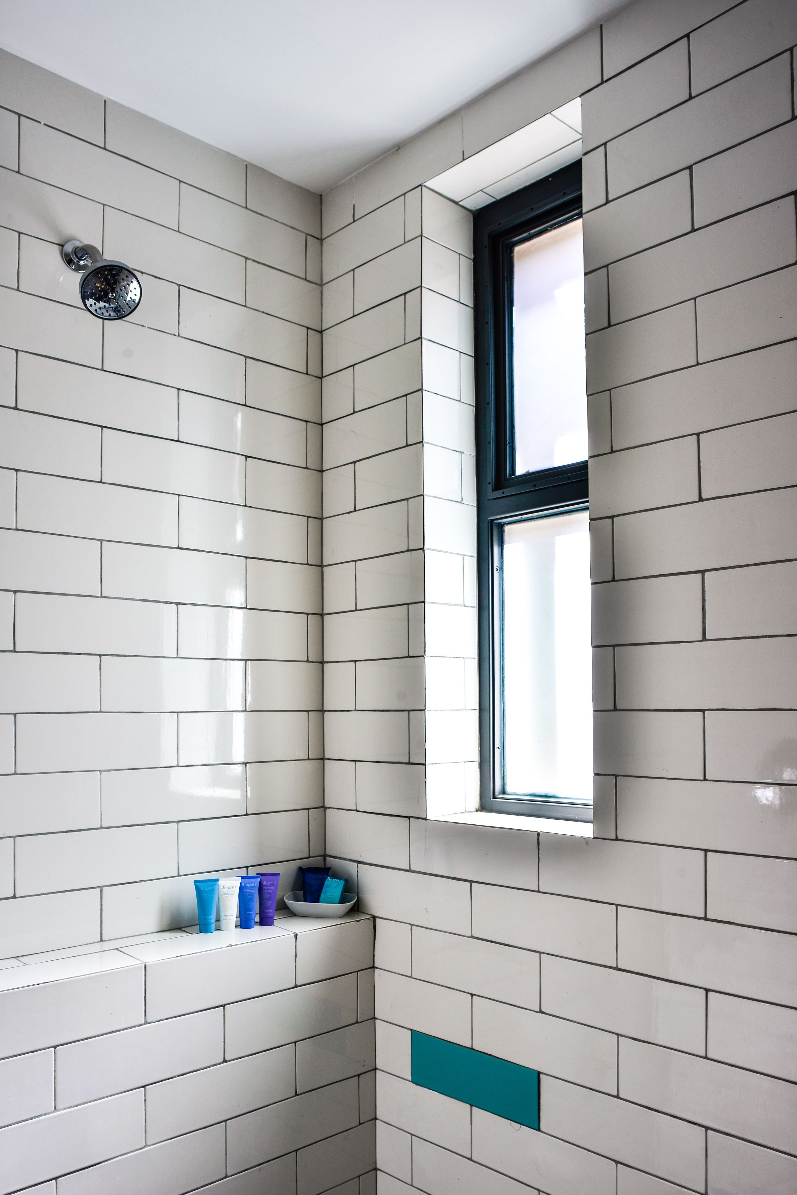 Een badkamer met witte betegelde wanden, een douchekop, een raam en een plank met toiletartikelen.