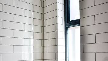 Een badkamer met witte betegelde wanden, een douchekop, een raam en een plank met toiletartikelen.