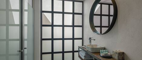 A modern bathroom with a large glass door, a round mirror, a sink, and a wooden shelf with towels and toiletries.