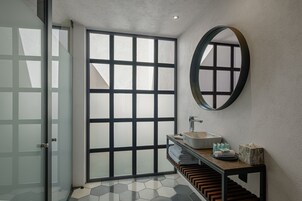 Un baño moderno con una amplia puerta de cristal, un espejo redondo, un lavamanos y una estantería de madera con toallas y artículos de aseo.