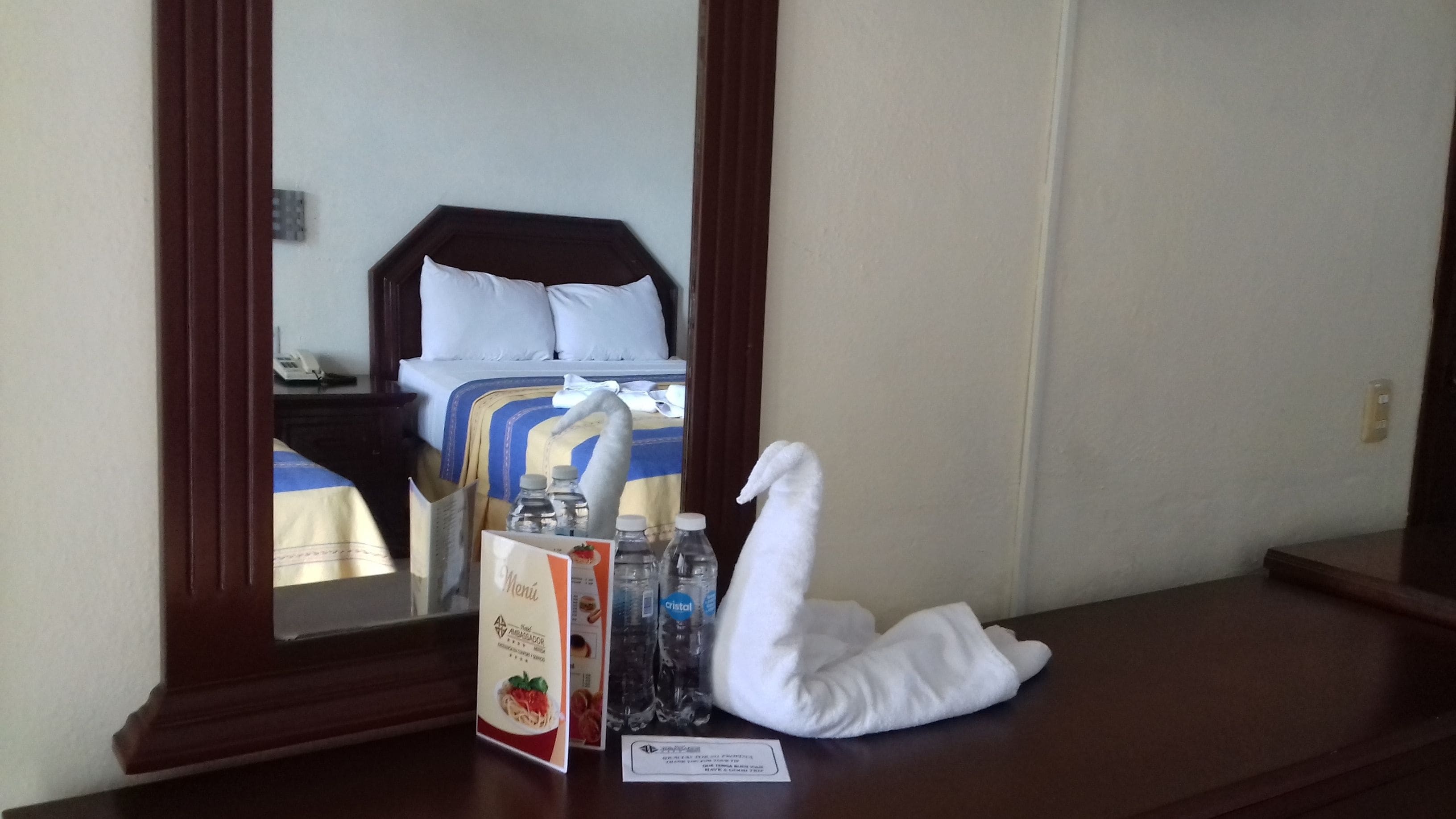 Ein Hotelzimmer mit einem Bett, einem Nachttisch aus Holz, einem Spiegel, einem schwanenförmigen Handtuch, einer Speisekarte und Wasserflaschen.