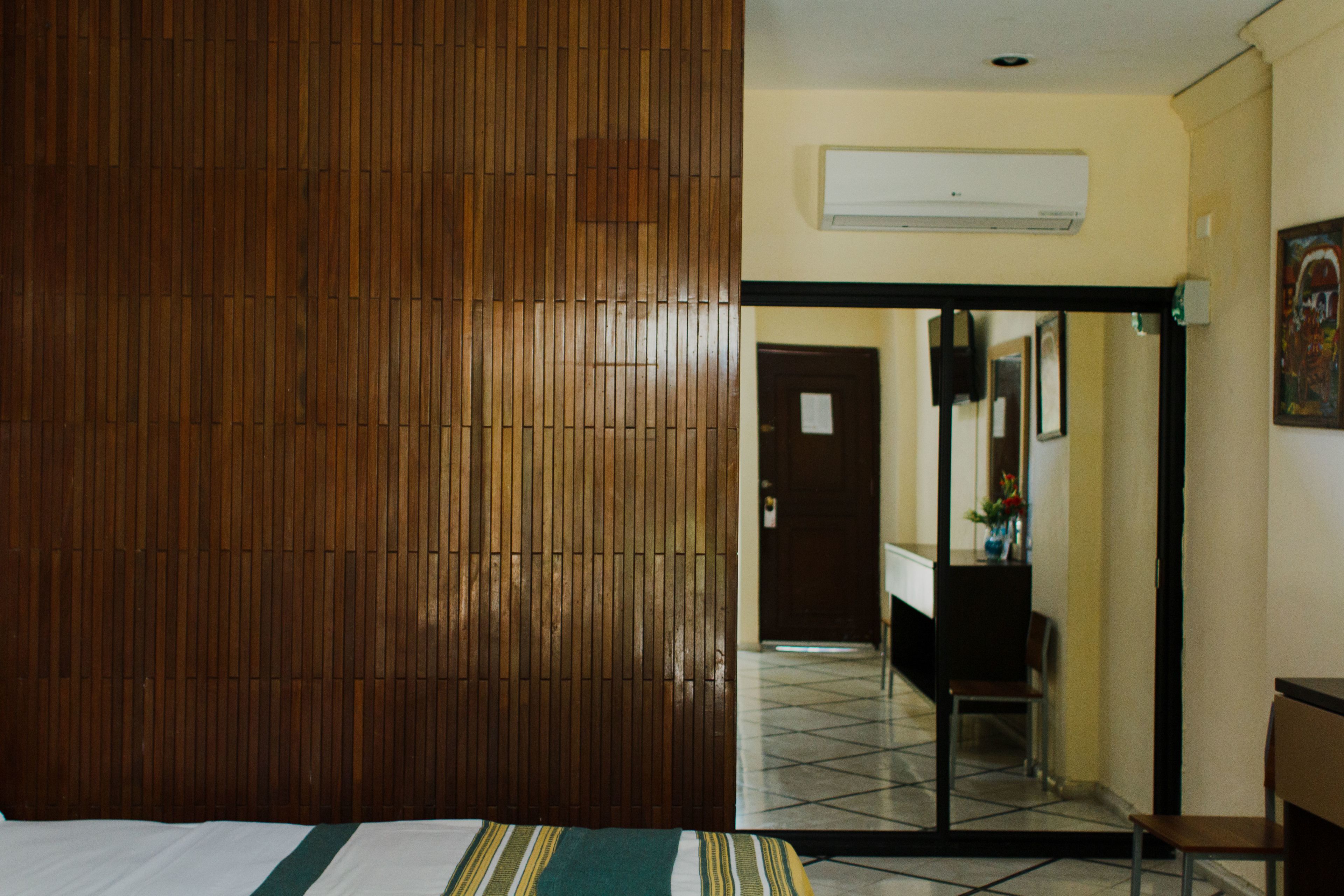 A hotel room with a wooden panel wall, a bed with a striped bedspread, a tiled floor, a desk with a chair, and a door leading to another room.