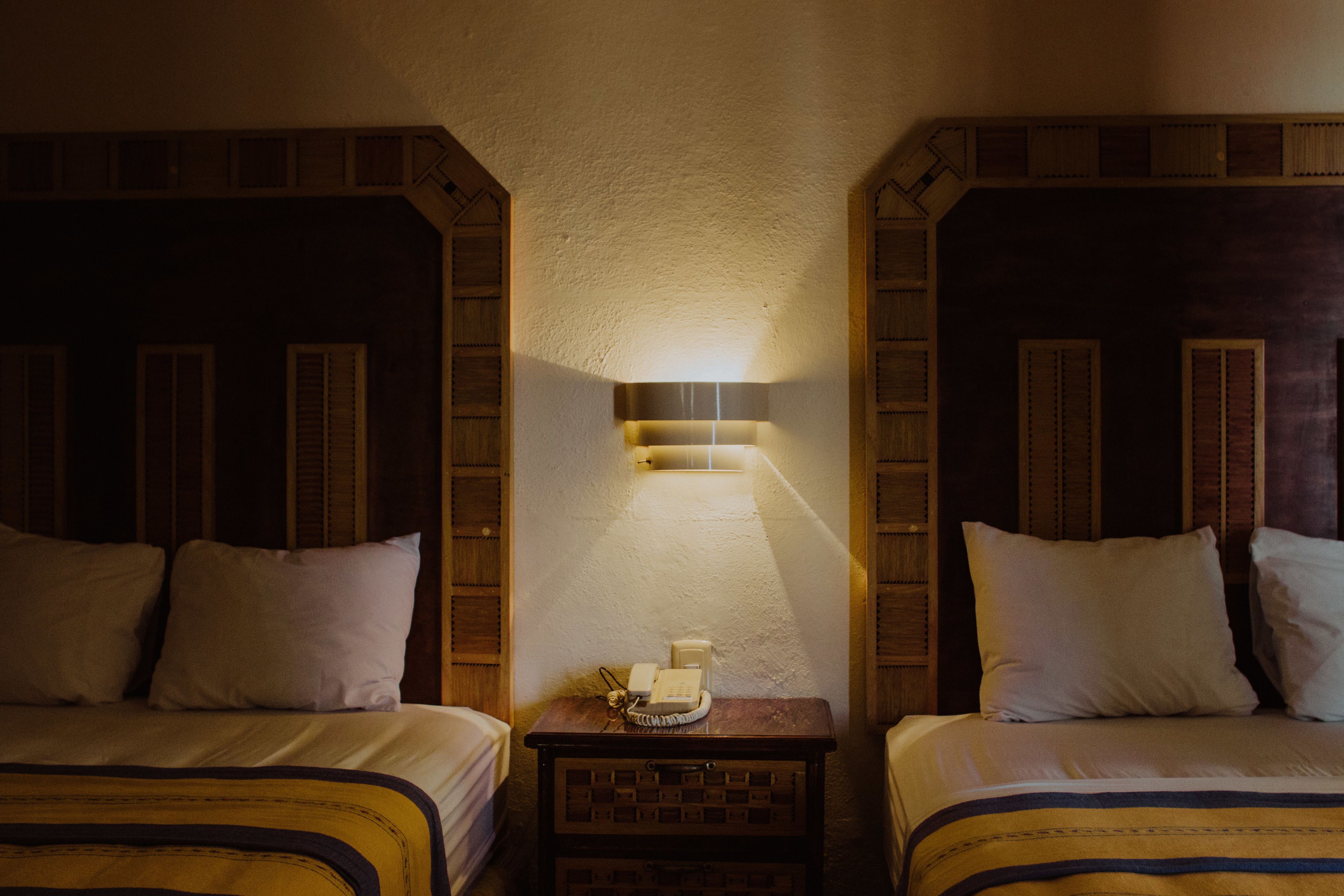A hotel room with two beds, a nightstand with a lamp, and a wall-mounted telephone.