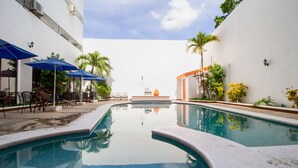 A swimming pool with a clear view of the sky, surrounded by chairs and umbrellas, and a building with a white facade.