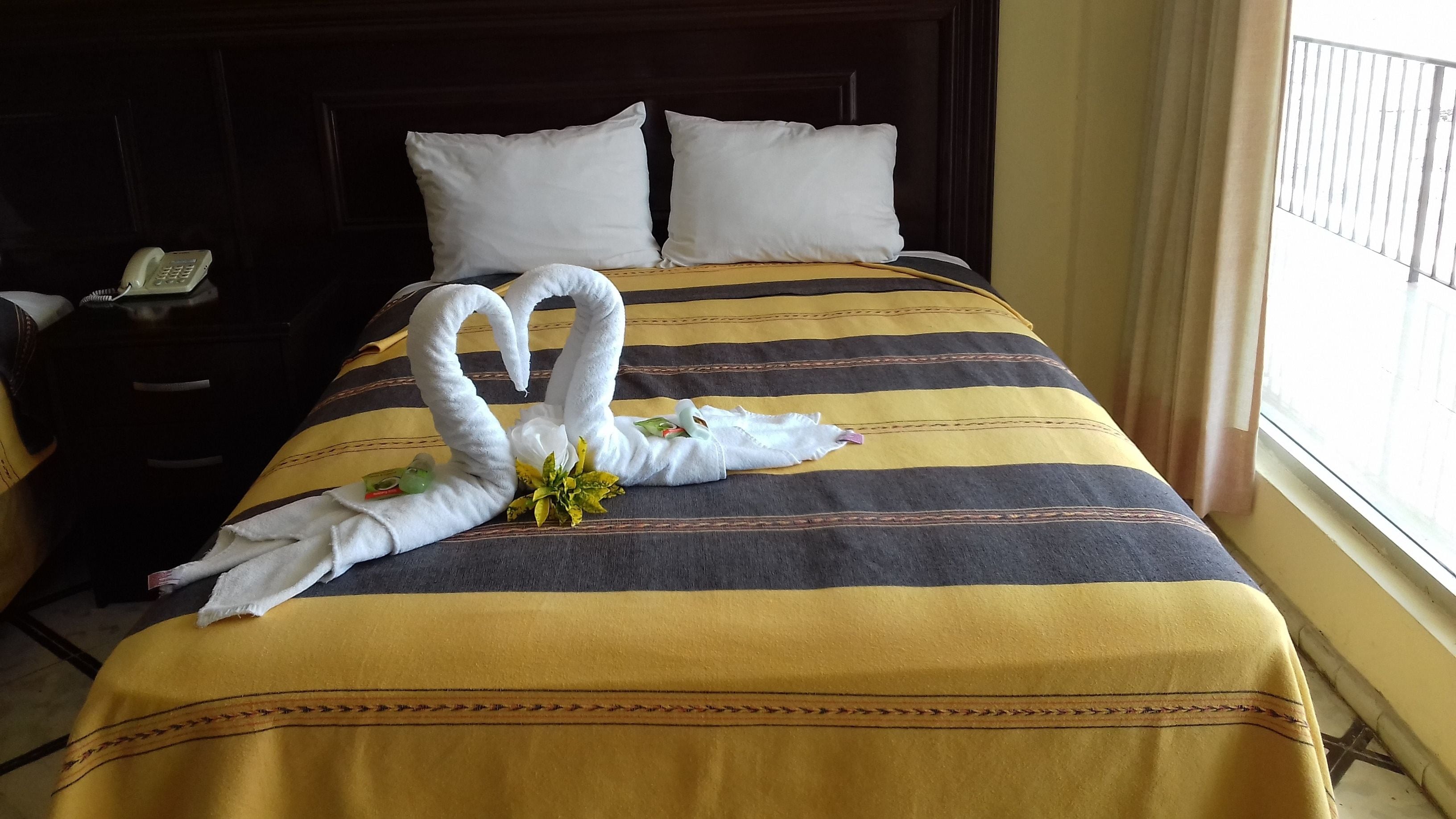 Une chambre d’hôtel avec un lit recouvert d’une couvre-lit à rayures, deux oreillers, une table de chevet avec un téléphone et une fenêtre avec des stores.
