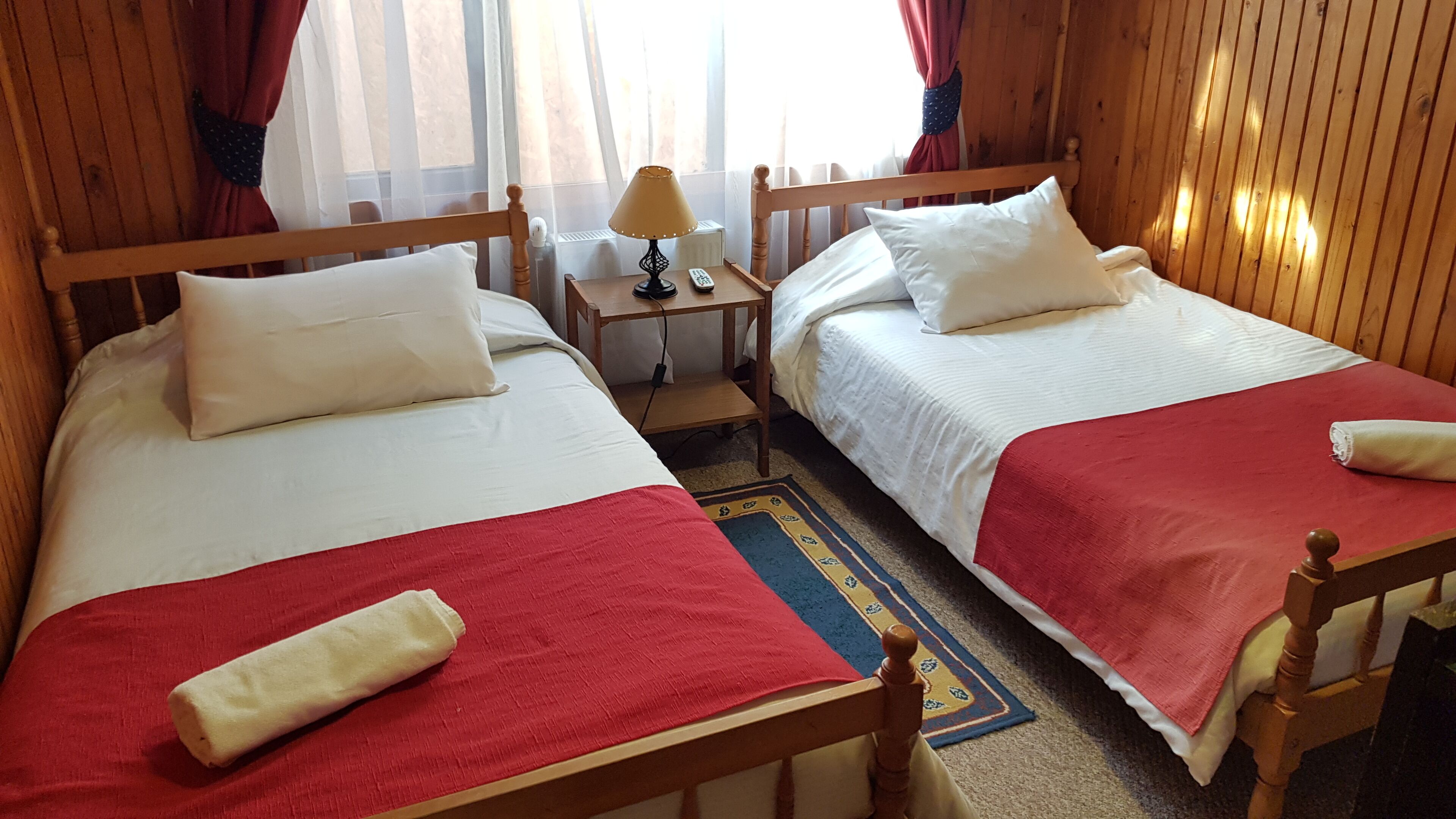 Una habitación doble con dos camas individuales, cada una con ropa blanca y mantas rojas. Hay una mesita de noche de madera con una lámpara y un control remoto entre las camas.
