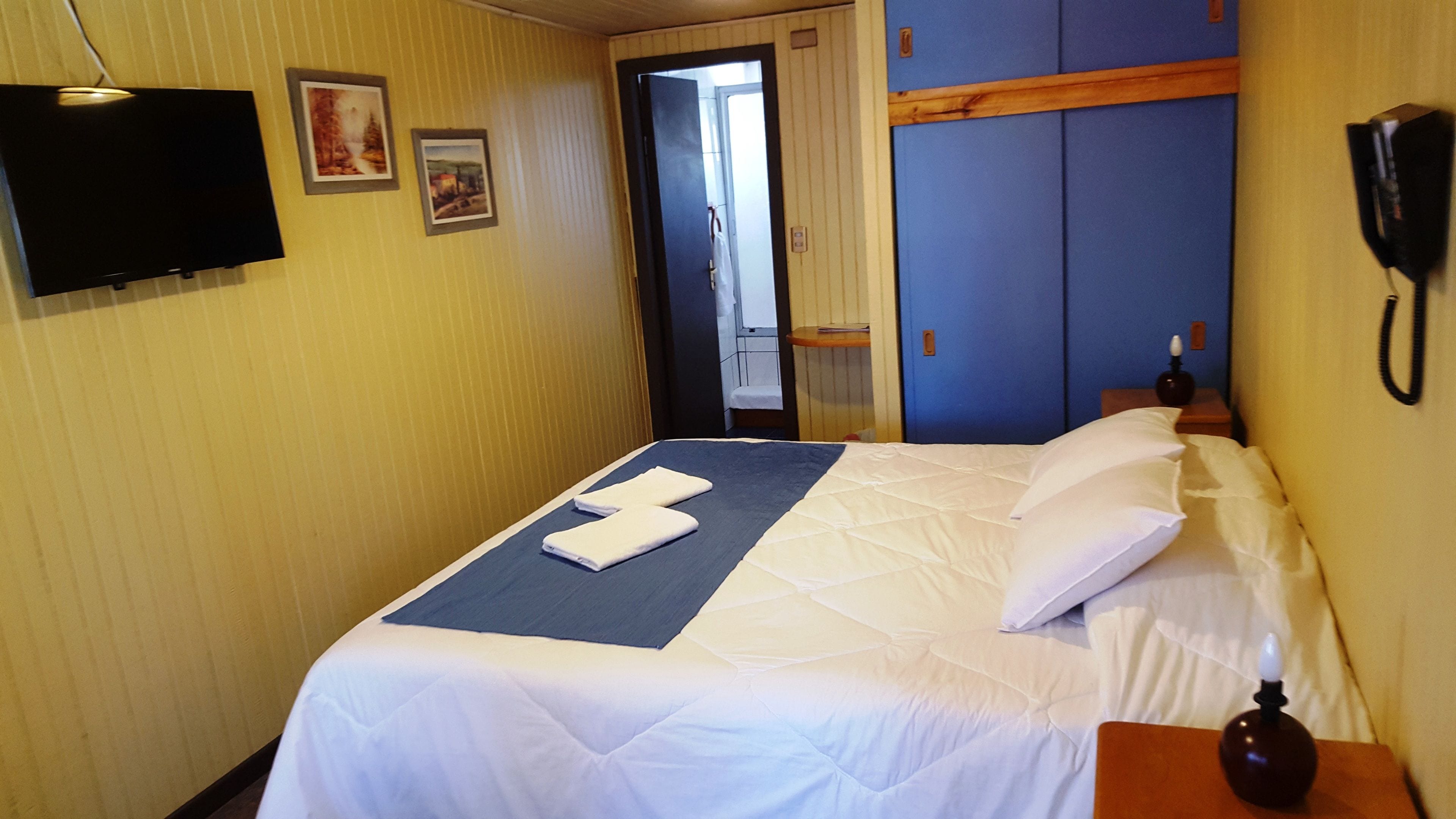 Quarto de hotel com uma cama, televisão de tela plana na parede e telefone na mesinha de cabeceira.