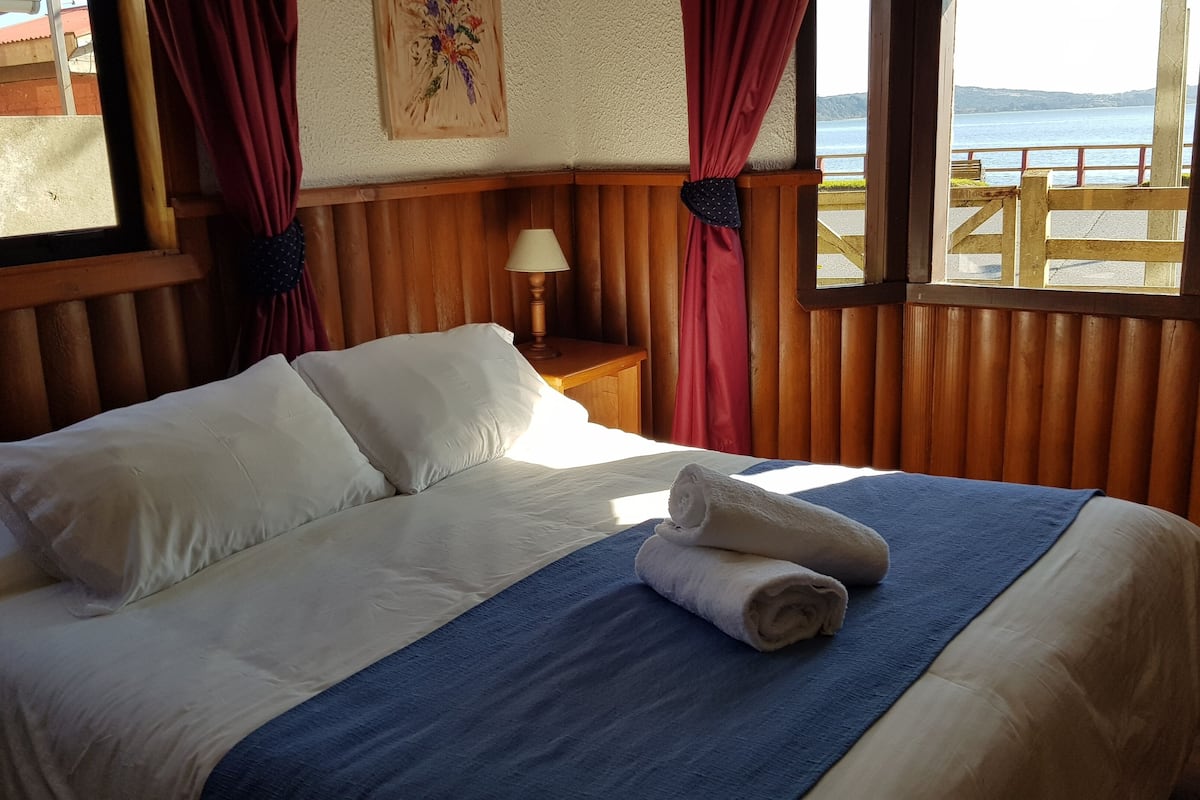 A bed with white linens and a blue blanket, a wooden headboard, a nightstand with a lamp, and a view of a body of water through a window with wooden railings.