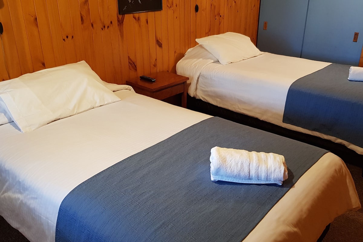 Two single beds with white linens and blue bedspreads, a wooden wall, and a small wooden nightstand with a remote control.
