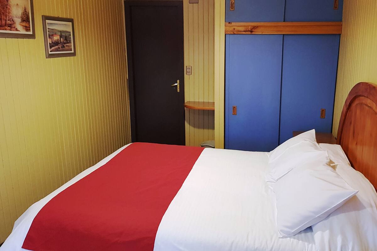 A single bed with a red and white bedspread, a wooden headboard, and a blue wardrobe in the background.