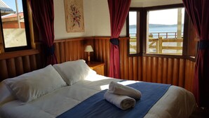 A neatly made bed with a blue blanket and rolled towels, wooden paneling on the walls, a nightstand with a lamp, and a window with a view of the sea. - Hostal Vista al Mar (Ancud)