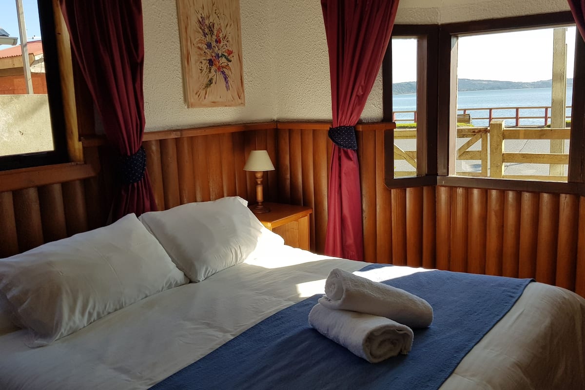 A neatly made bed with a blue blanket and rolled towels, wooden paneling on the walls, a nightstand with a lamp, and a window with a view of the sea.