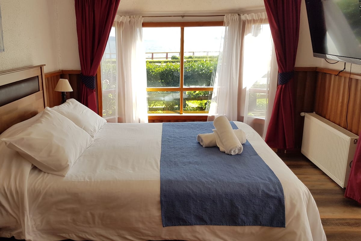 A neatly made bed with white and blue linens, a wooden headboard, and a view of greenery through a window with red curtains.