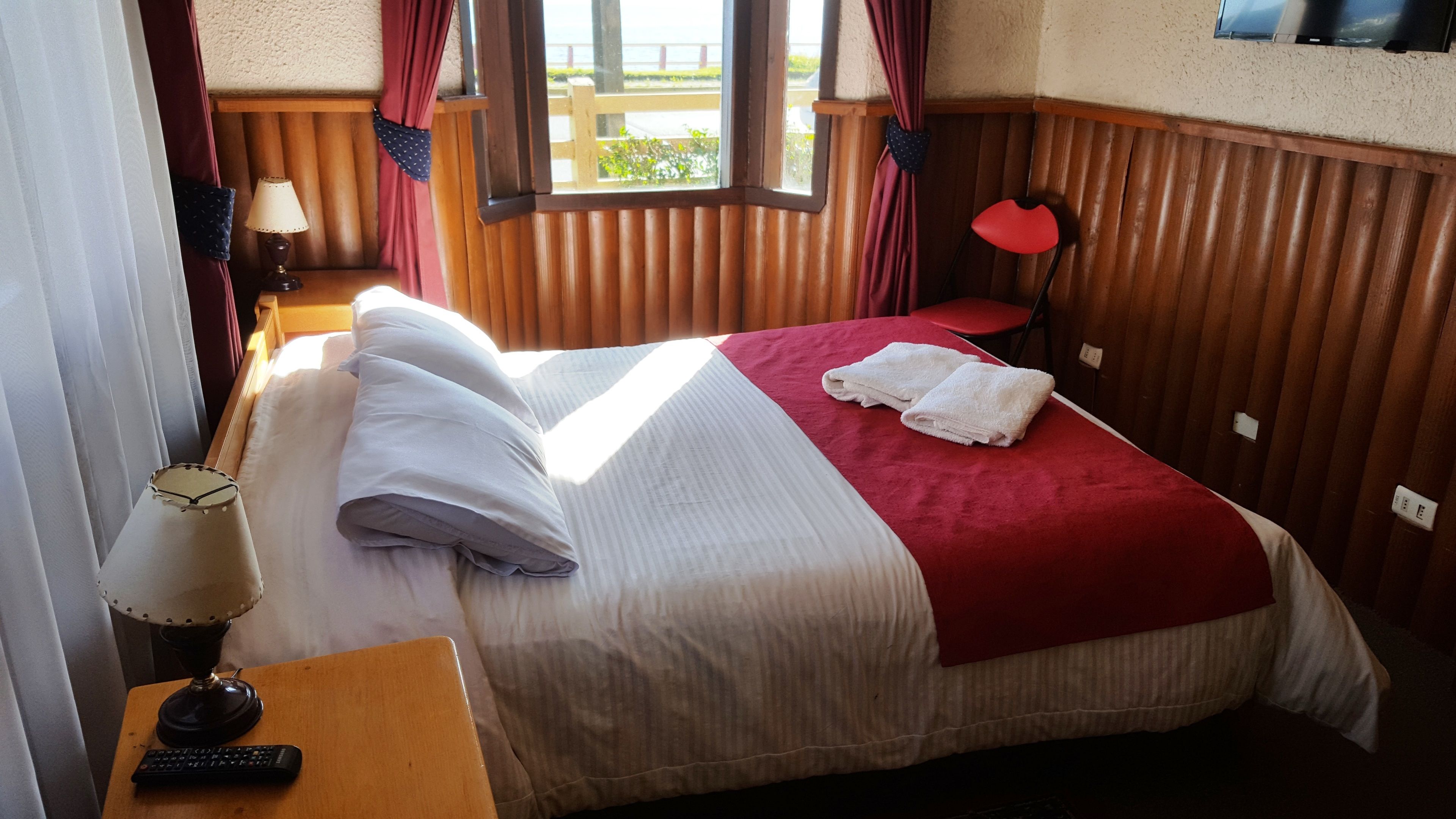 Una habitación de hotel con una cama, una mesita de noche, una lámpara y un ventanal con cortinas.