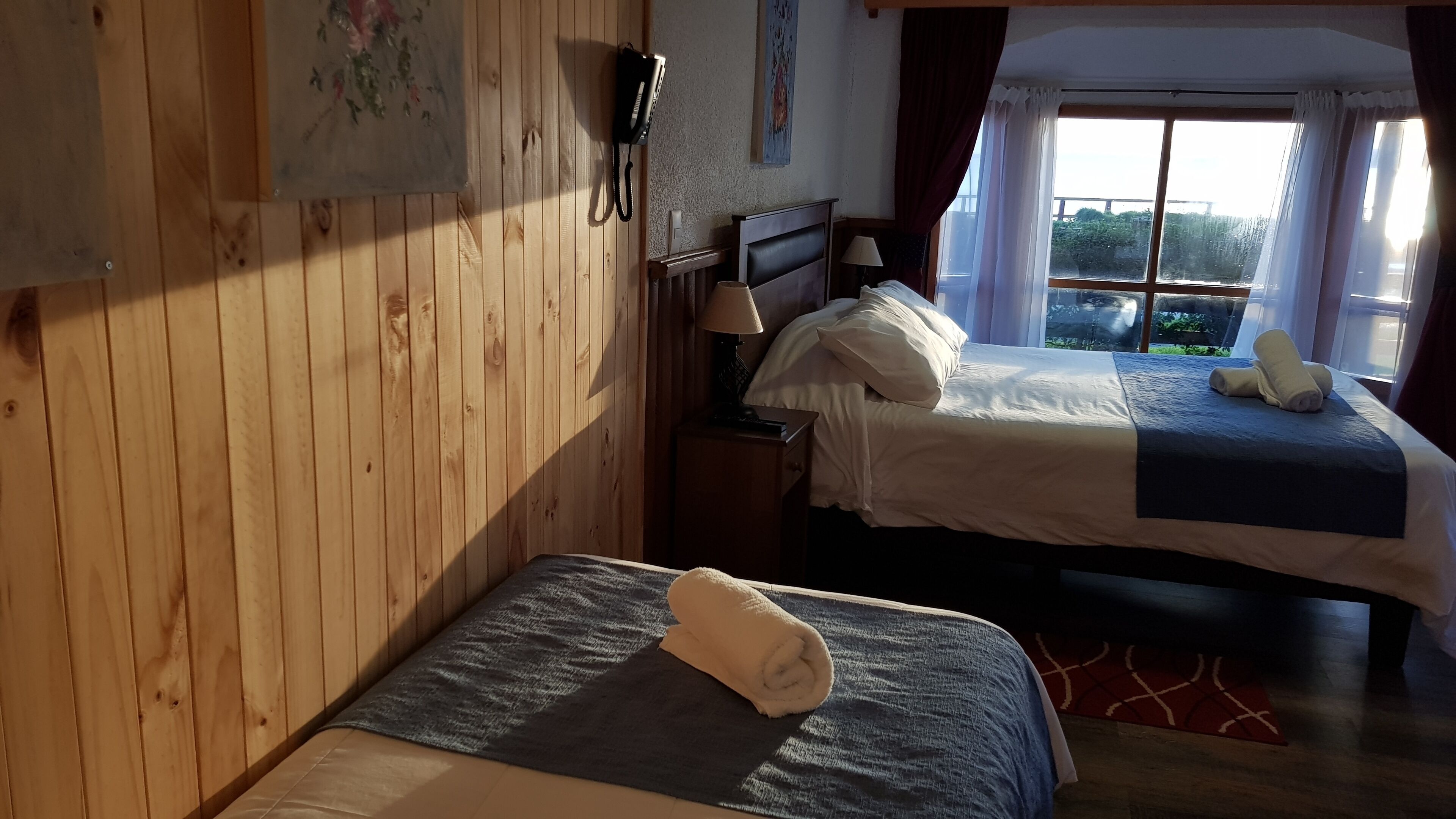 Un dormitorio con dos camas, paredes de madera, un cuadro, un teléfono y una ventana con cortinas.