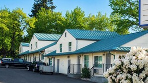 A row of motel buildings with green roofs, a parking area with vehicles, and a large white flowering bush. - Budget Lodge by OYO Portland West (Portland)