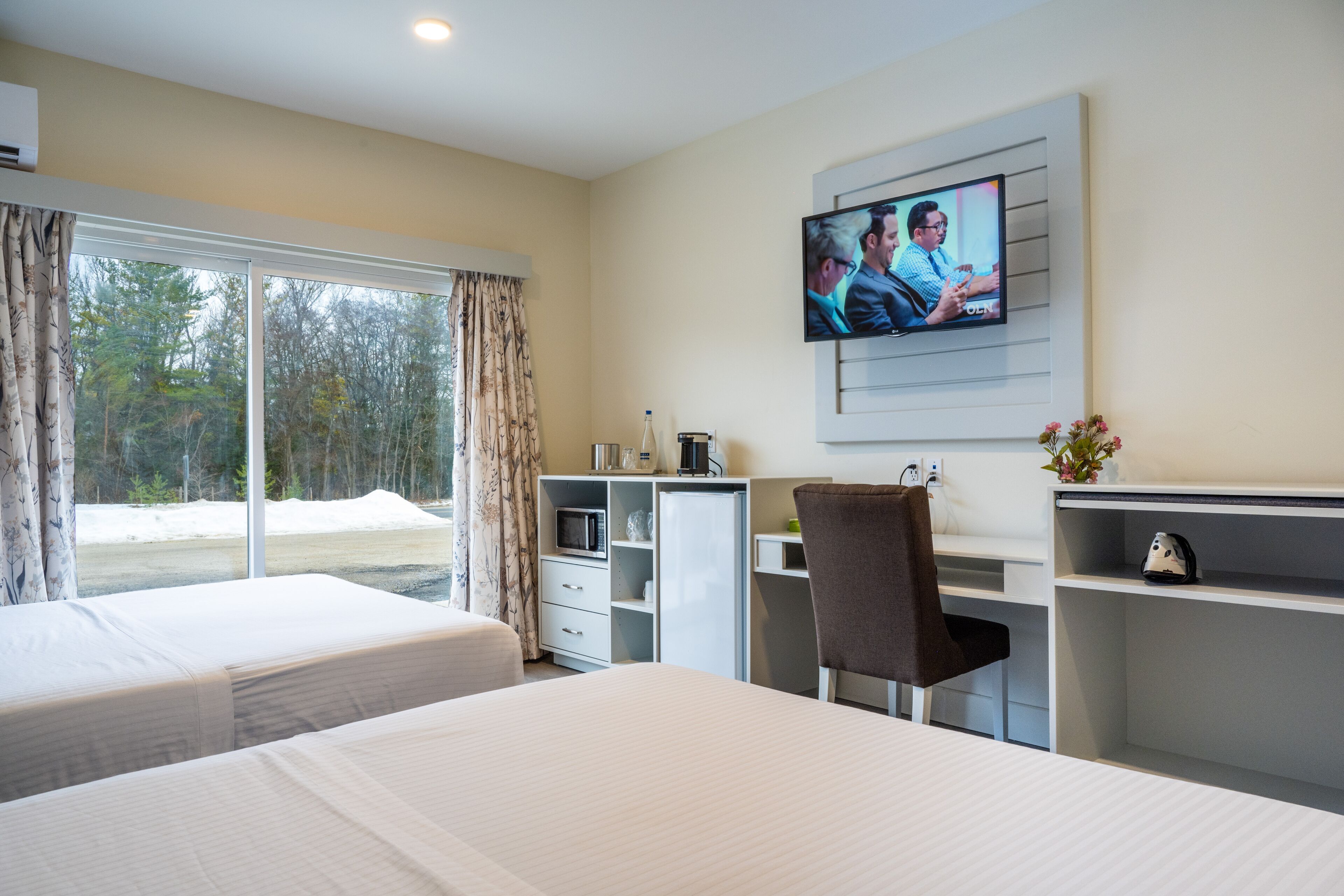 A hotel room with two beds, a desk, a TV, and a view of trees through the window.