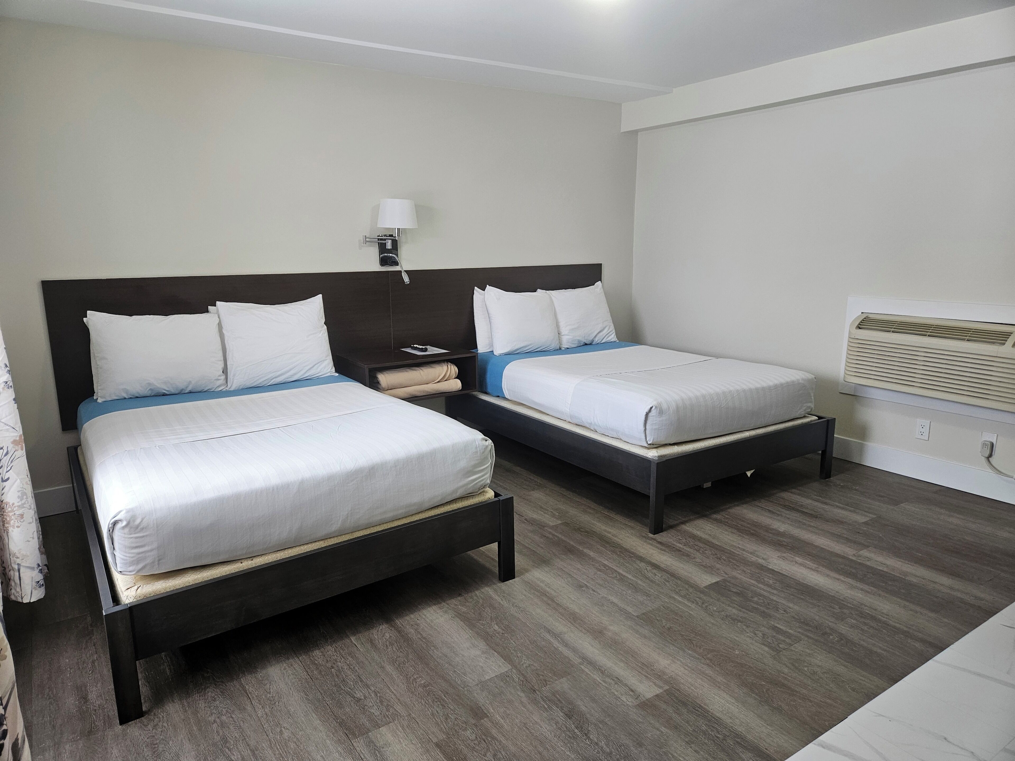 Habitación de hotel con dos camas, una mesita de noche, una lámpara y un aire acondicionado instalado en la pared.