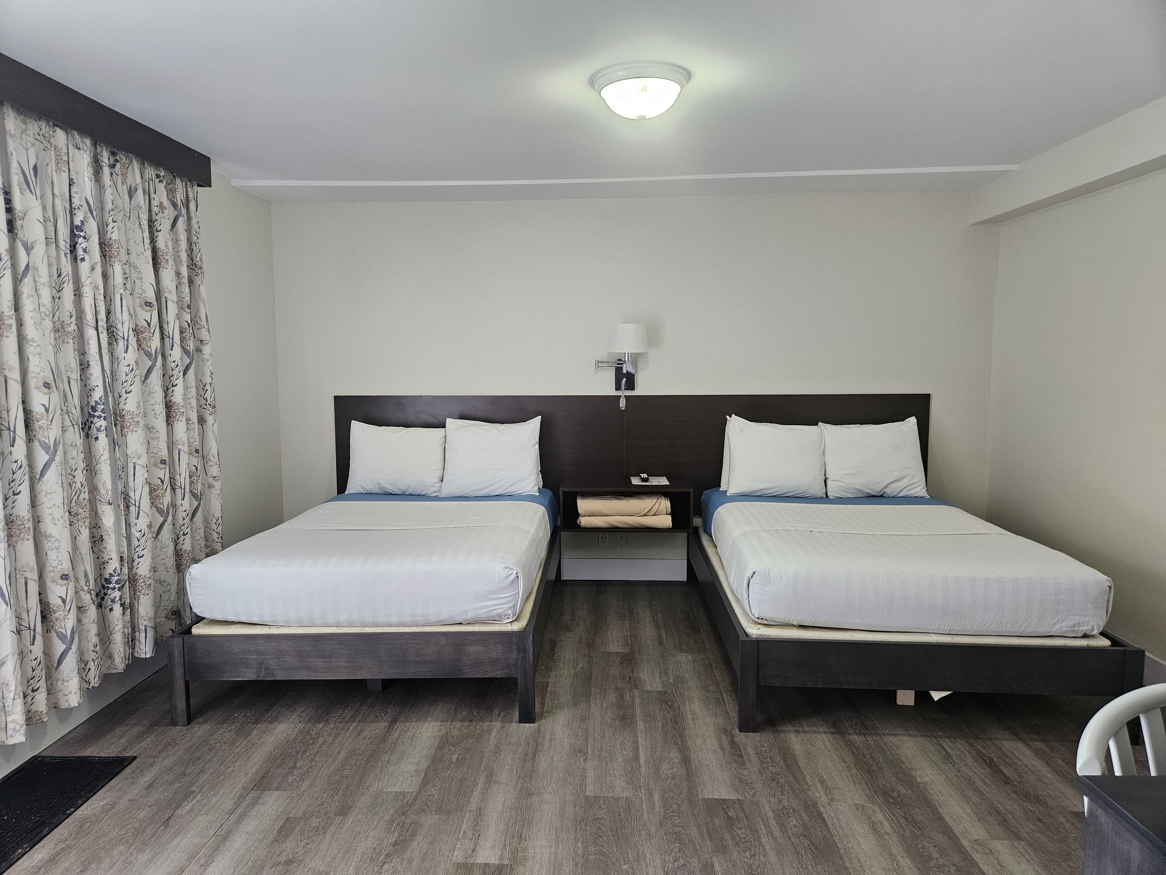 Ein Hotelzimmer mit zwei Betten, einem Nachttisch, einer Lampe und einem Fenster mit Vorhängen.