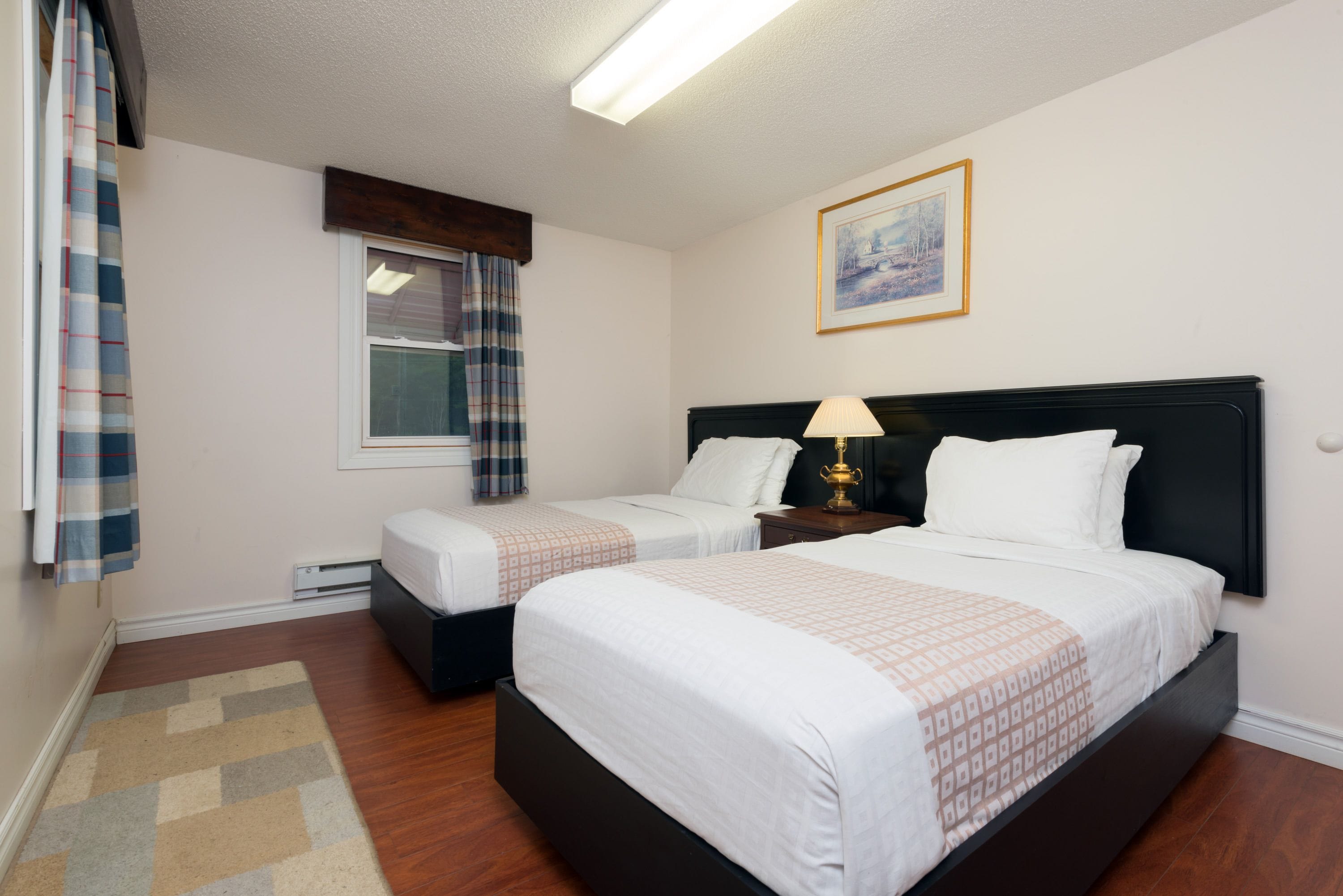A hotel room with two beds, a window with curtains, a framed picture on the wall, and a bedside lamp.