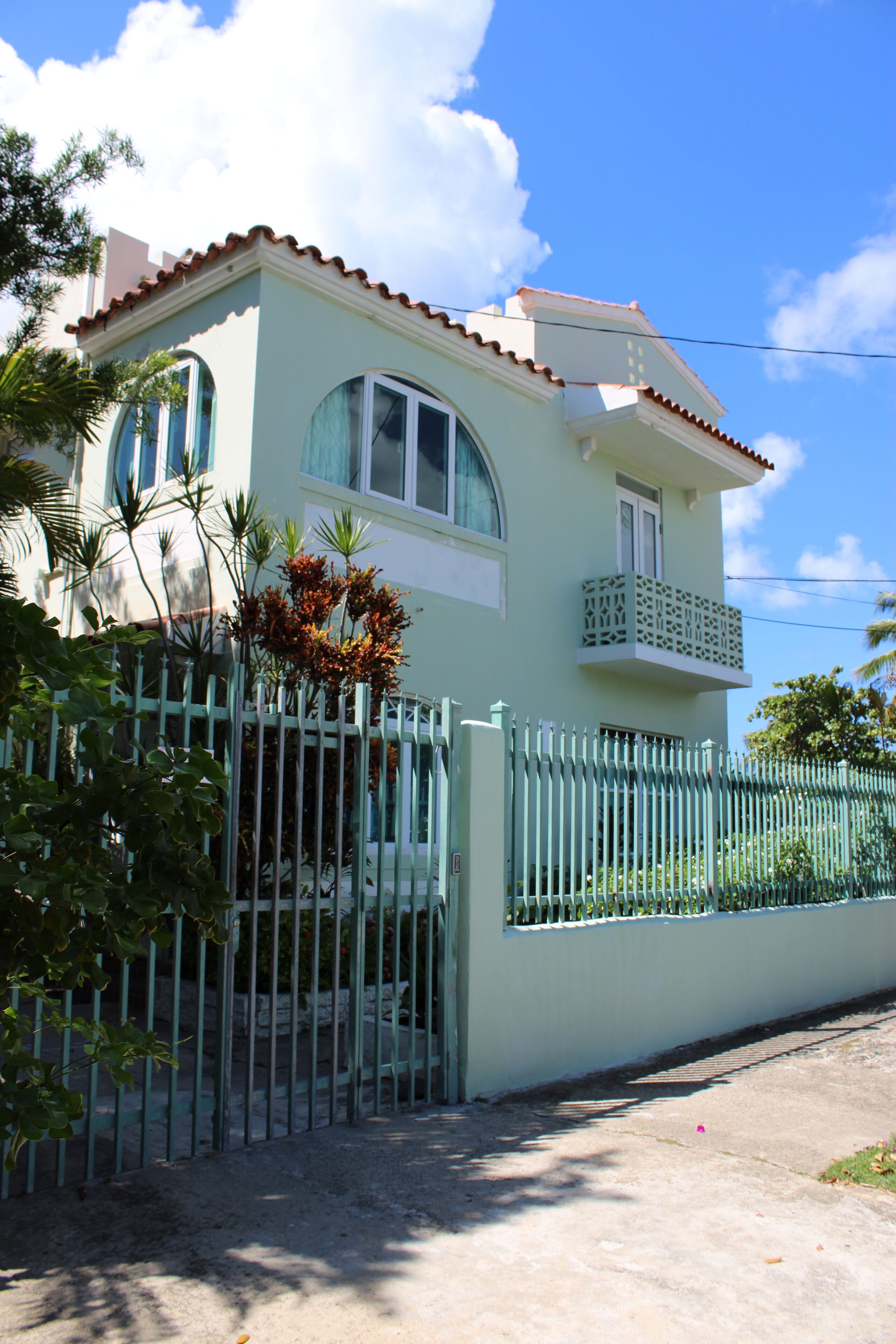 Una casa de dos pisos con fachada verde clara, un balcón blanco y una valla de piquetes blanca.