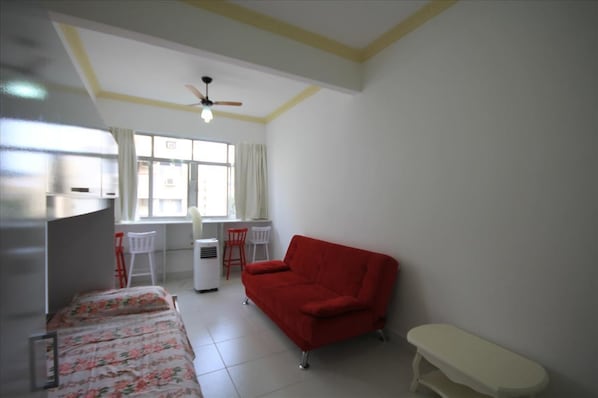 A room with a bed, a red sofa, a desk with a chair, a white bench, and a ceiling fan. - Bernardes 808 (Rio de Janeiro)