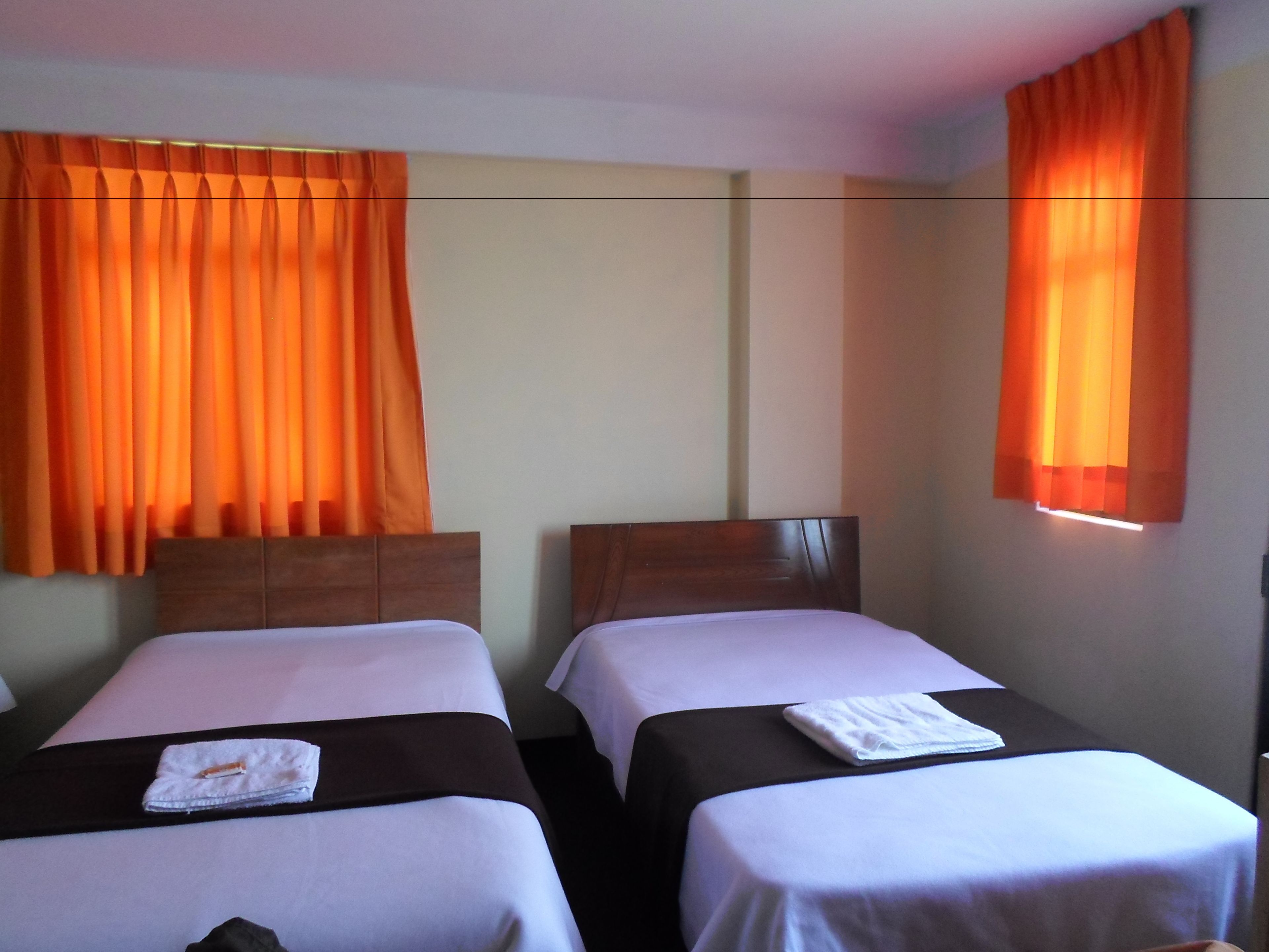 A hotel room with two single beds, each with a wooden headboard, white bedding, and an orange curtain.