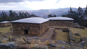 Un edificio de piedra con techo plano, rodeado por un muro de piedra y una zona cubierta de césped.