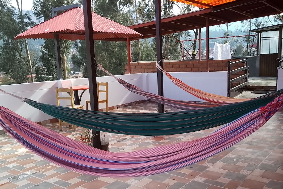 A hammock on a tiled patio with a red roof and a view of trees and buildings in the background.