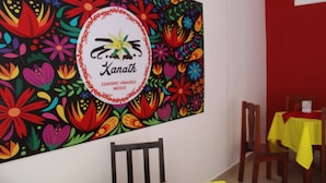 Een kleurrijke bloemenmuurschildering met een logo en tekst op een muur in een eetgedeelte met houten stoelen en een gele tafelkleed.