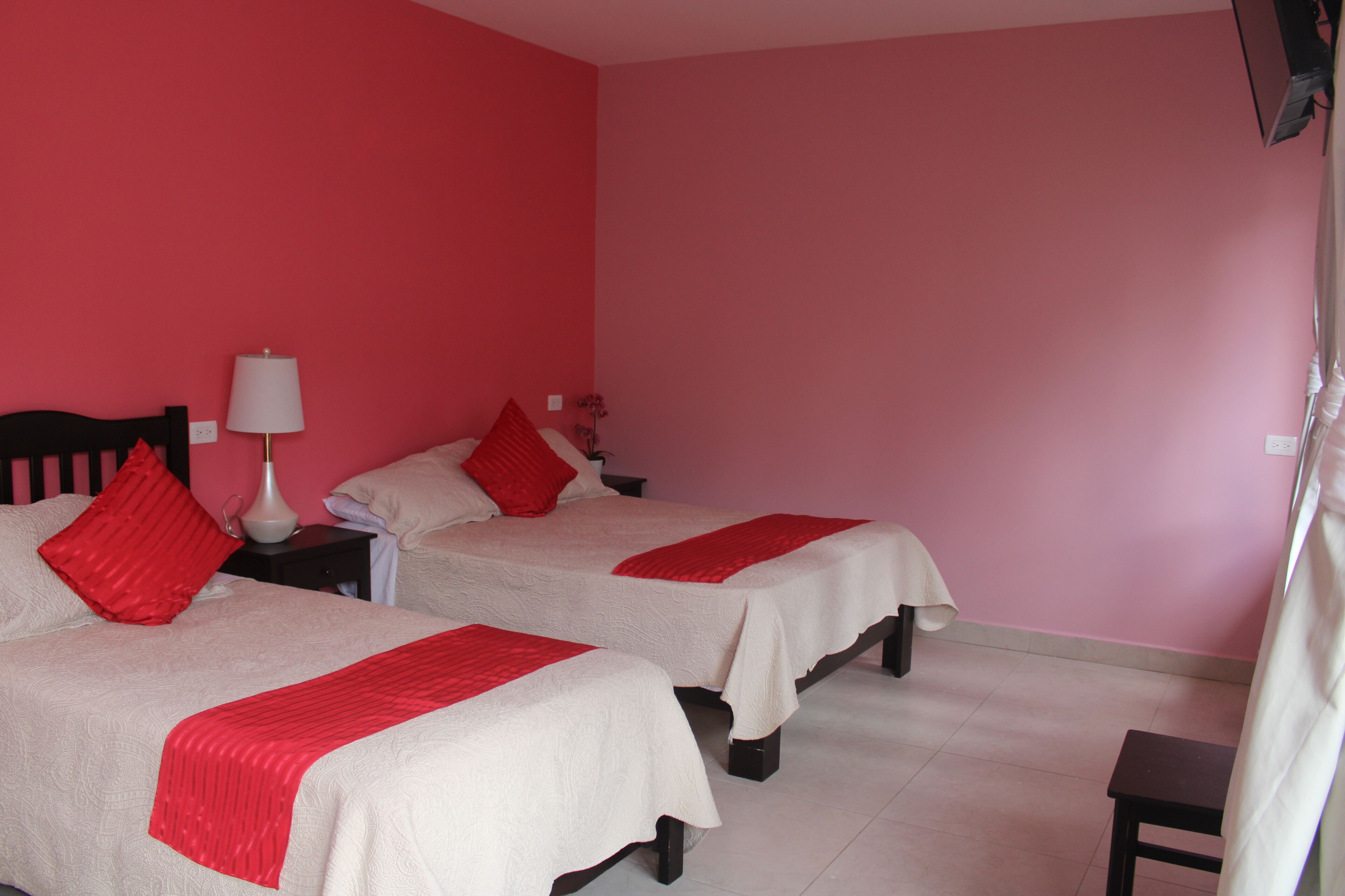 A room with two beds, each with a red pillow and a white bedspread. There is a nightstand with a lamp between the beds.