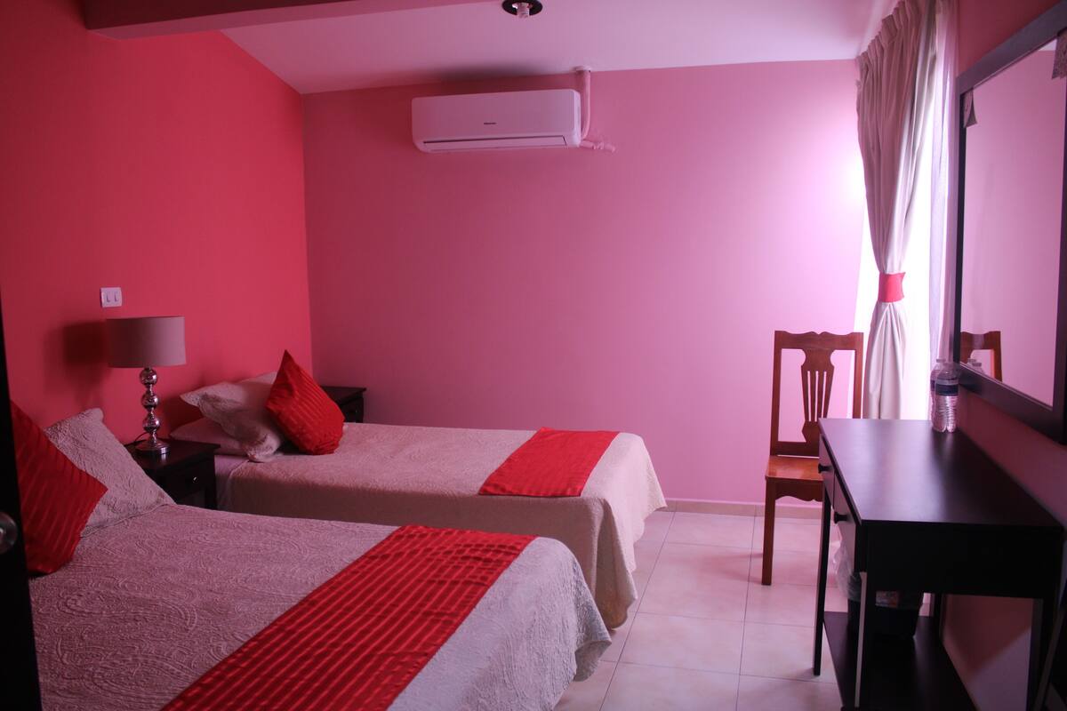 Ein Zimmer mit zwei Betten, einem Schreibtisch, einem Stuhl, einer Lampe und einer Klimaanlage.