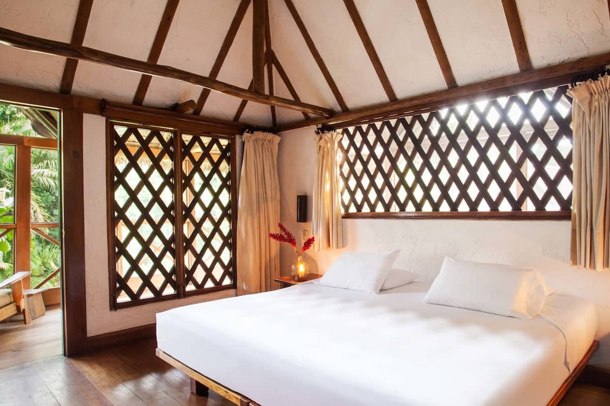A bedroom with a large bed, wooden beams, and a view through a lattice window.