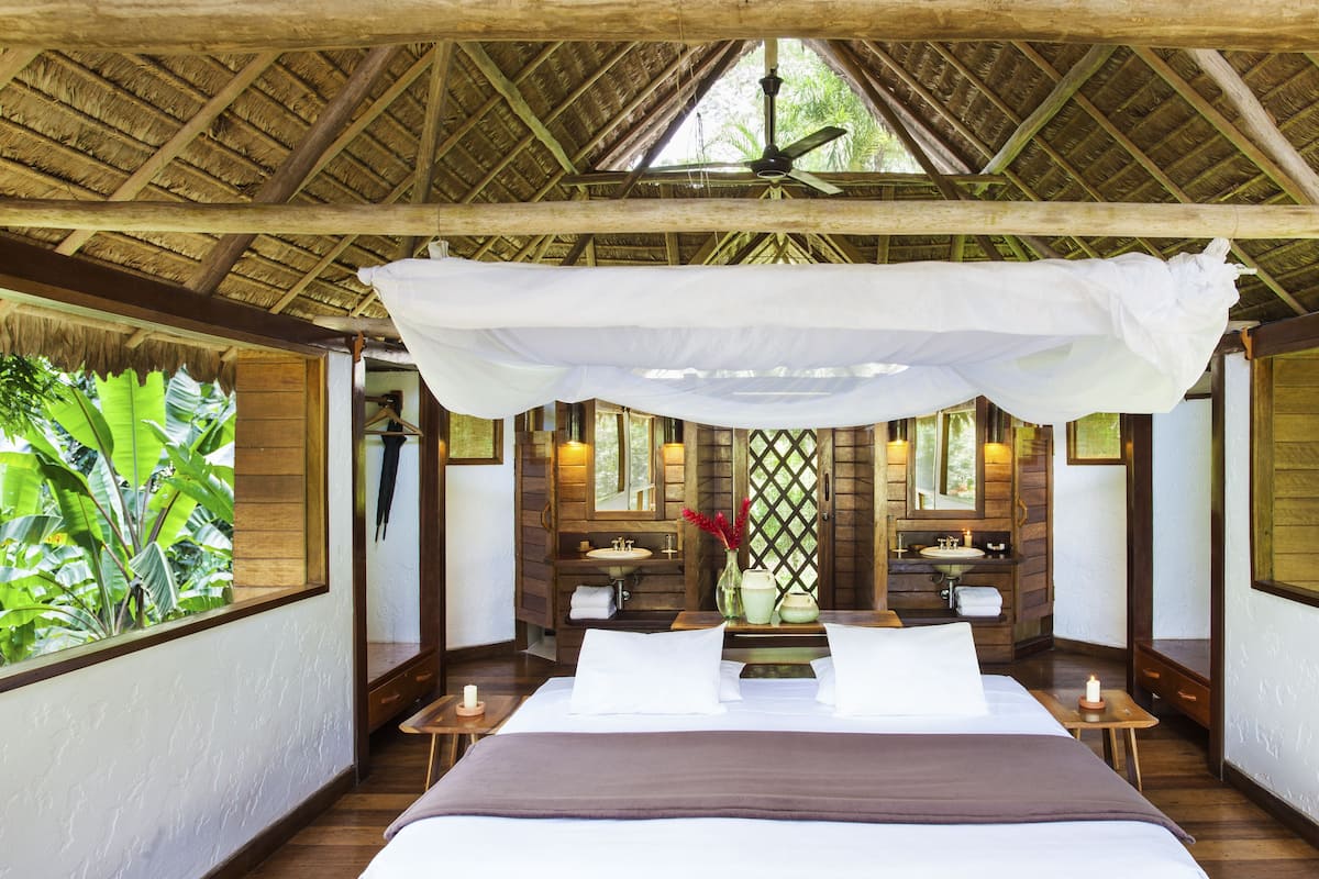 A room with a bed, a ceiling fan, a window with a view of greenery, a mirror, and a small table with a candle.