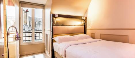A hotel room with a bed, a window with a view of buildings, and a door with a towel rack.