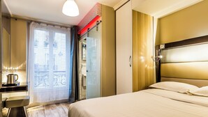 A hotel room with a bed, a bedside table with a lamp, a window with a view, and a door leading to a bathroom. - District Republique (Paris)