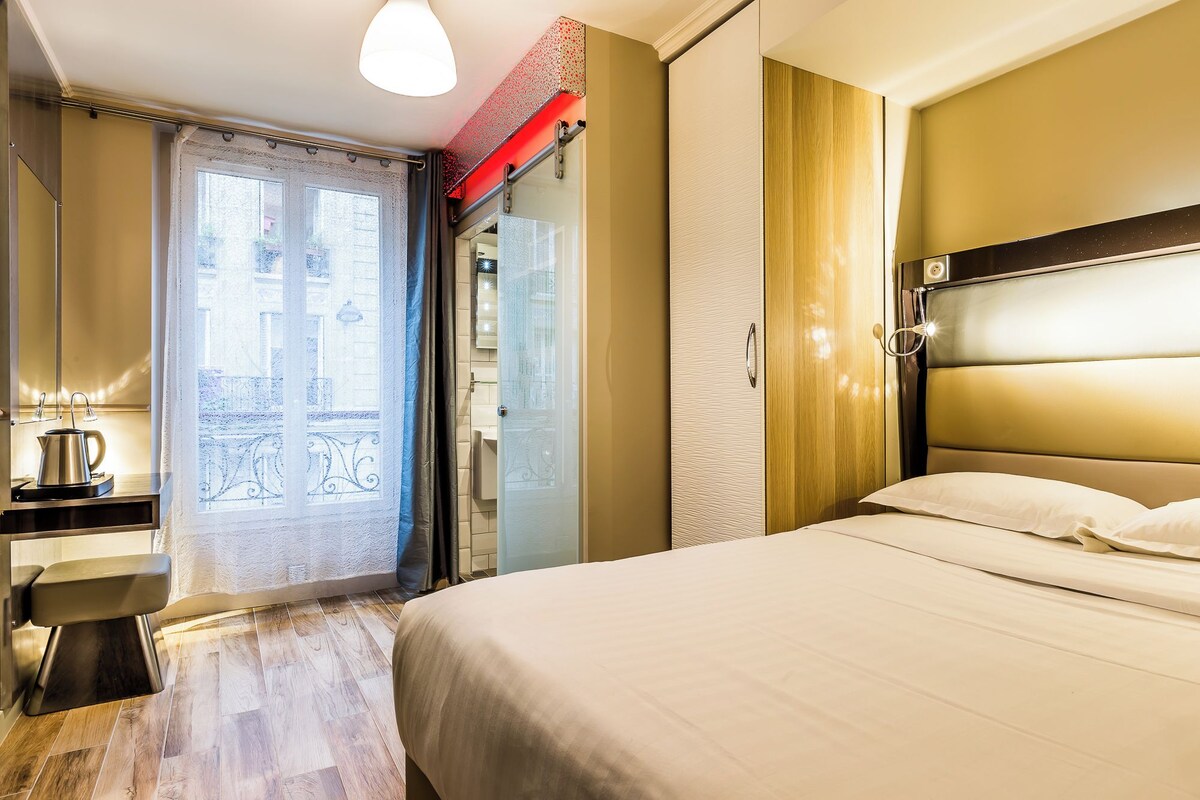 A hotel room with a bed, a bedside table with a lamp, a window with a view, and a door leading to a bathroom.